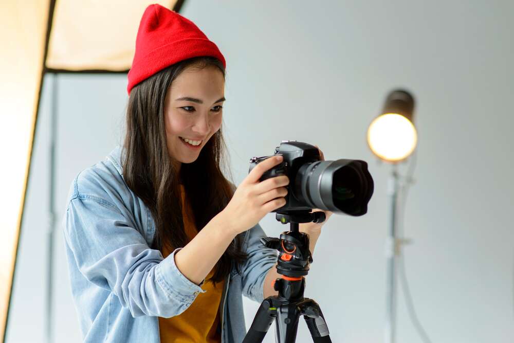 A young woman wearing a red beanie and a light blue overshirt smiles as she adjusts a professional camera mounted on a tripod inside a studio.