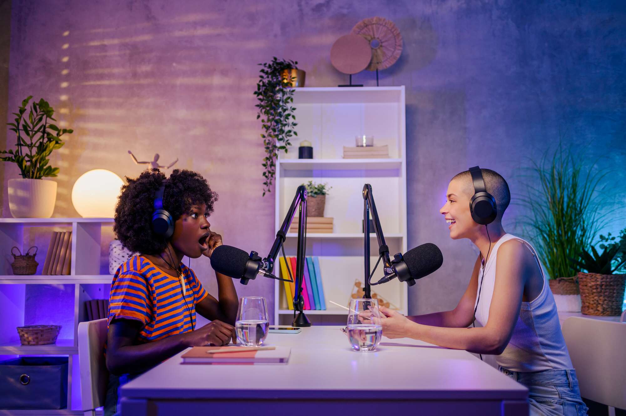 Two diverse women wearing headphones and speaking into microphones while recording a podcast at a table.
