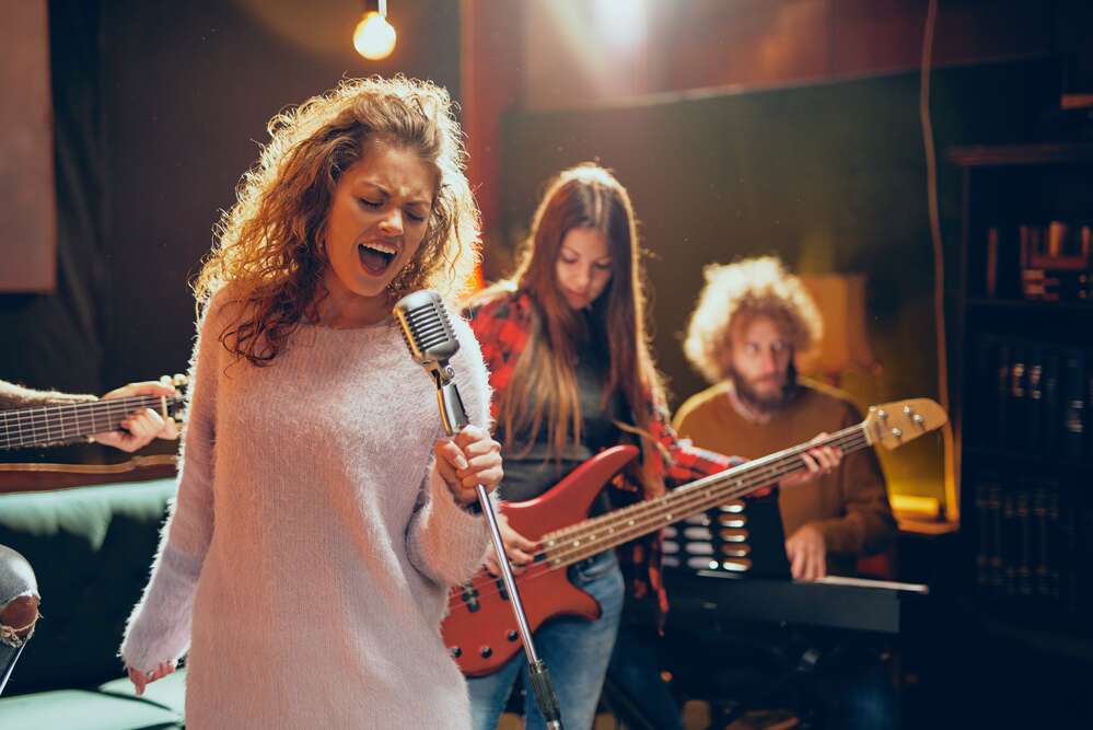 A rock band performing in a studio, with a woman singing into a vintage microphone in the foreground, and other band members playing a bass guitar and keyboard behind her.