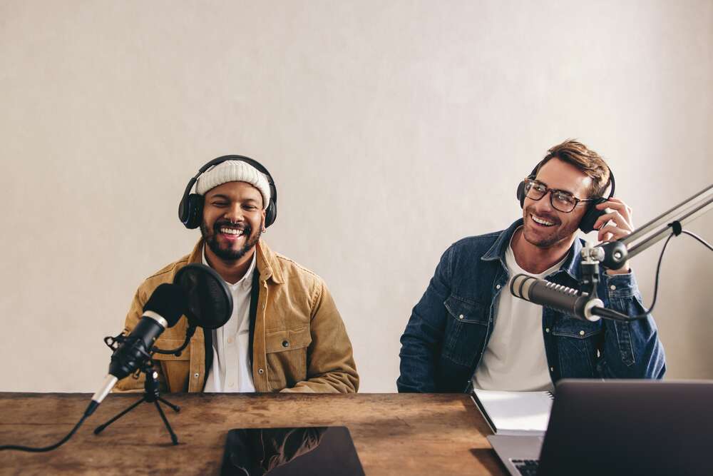 Two smiling men wearing headphones and recording a podcast in front of microphones.