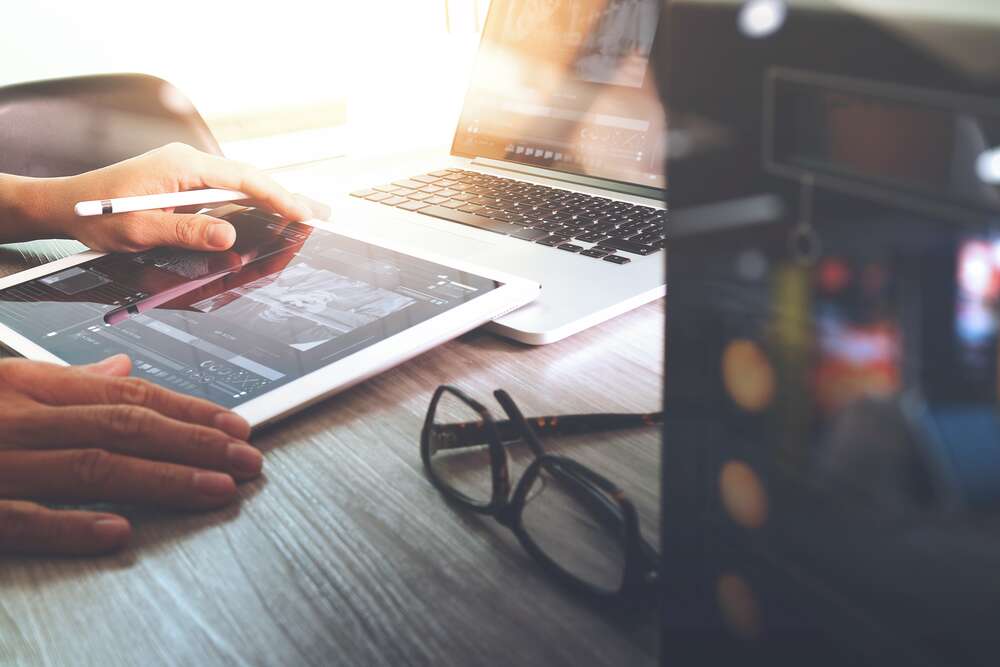 Close-up of a person using a stylus on a tablet next to a laptop and glasses on a wooden desk.