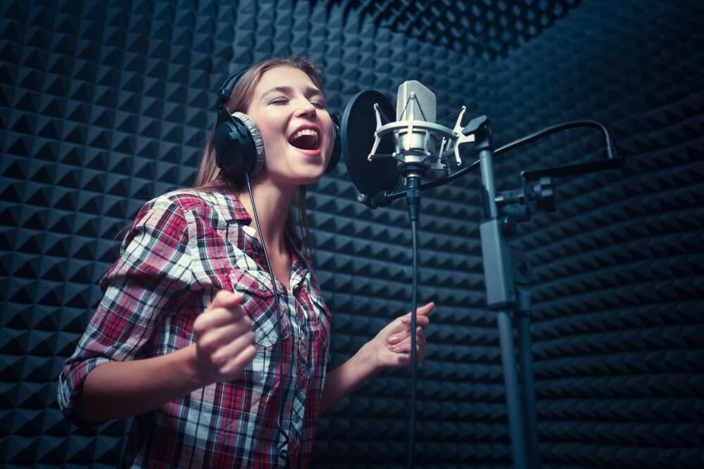 A young woman wearing headphones sings passionately into a studio microphone in a recording booth lined with acoustic foam panels.