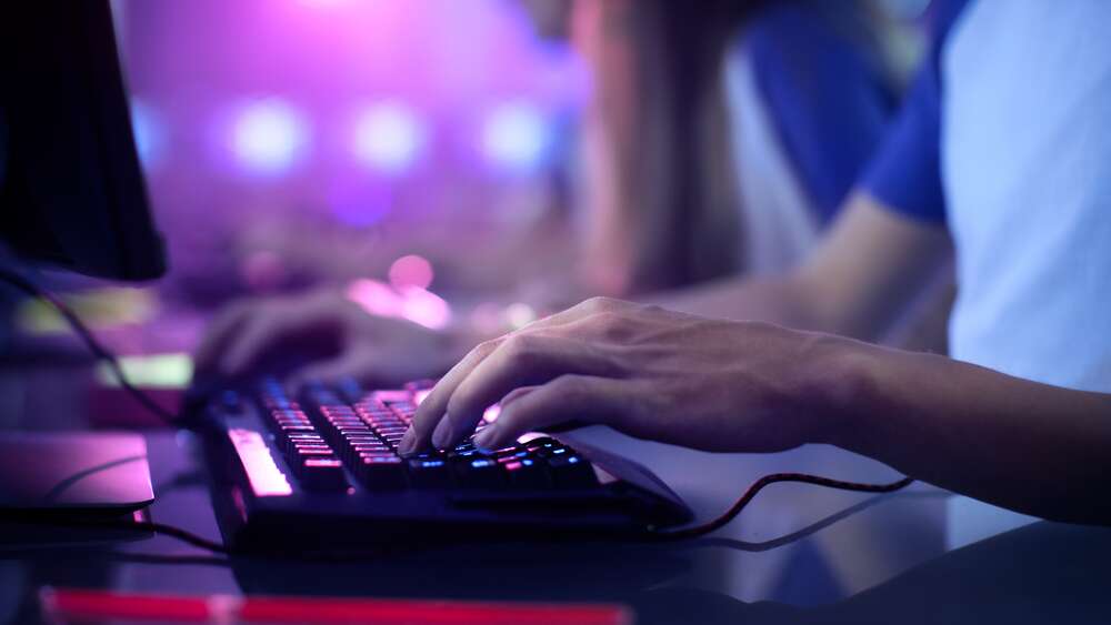 Close-up of a person's hands gaming on a backlit keyboard and mouse, with other players blurred in the background.
