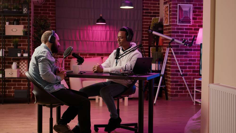 Two men wearing headphones sit across from each other at a desk in a podcast studio, speaking into microphones.