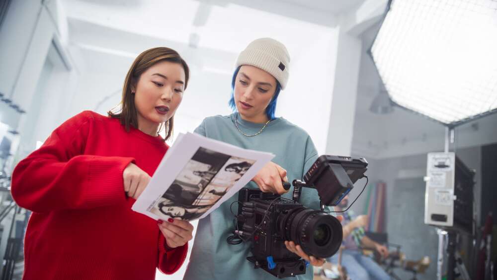 Two women collaborating on a video shoot, with one holding storyboard sketches and the other holding a professional camera in a studio setting.