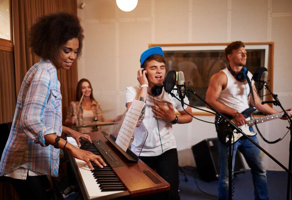 A band rehearses in a recording studio, featuring a keyboardist, a vocalist with headphones, a guitarist, and a drummer in the background.