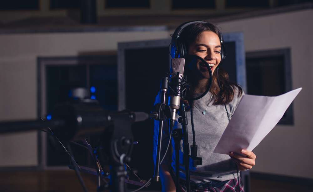A young woman wearing headphones and smiling while singing into a studio microphone and holding sheet music.